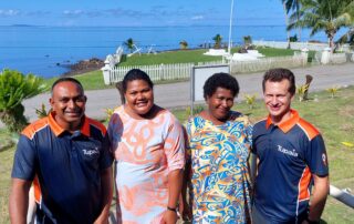 Four people, Shakti Gounder (Tamanu), Talei Qalo (CHW Wainaloka), Salai Murray (CHW Toki) and Geoff Fisher (Tamanu) stand in front of a grassy area leading to the ocean. They are all smiling.