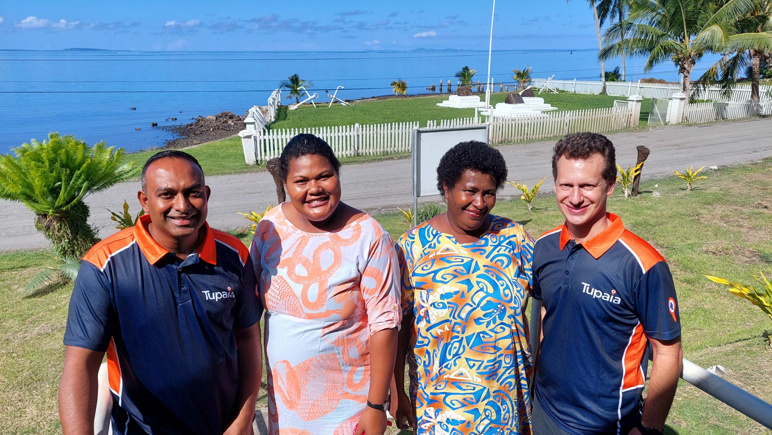 Four people, Shakti Gounder (Tamanu), Talei Qalo (CHW Wainaloka), Salai Murray (CHW Toki) and Geoff Fisher (Tamanu) stand in front of a grassy area leading to the ocean. They are all smiling.